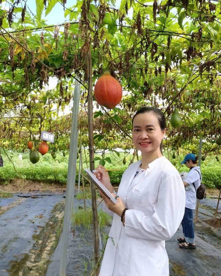 Nguyễn Hoa mặc áo blouse trắng, cầm sổ ghi chép dưới giàn gấc trong buổi thực tế vườn dược liệu